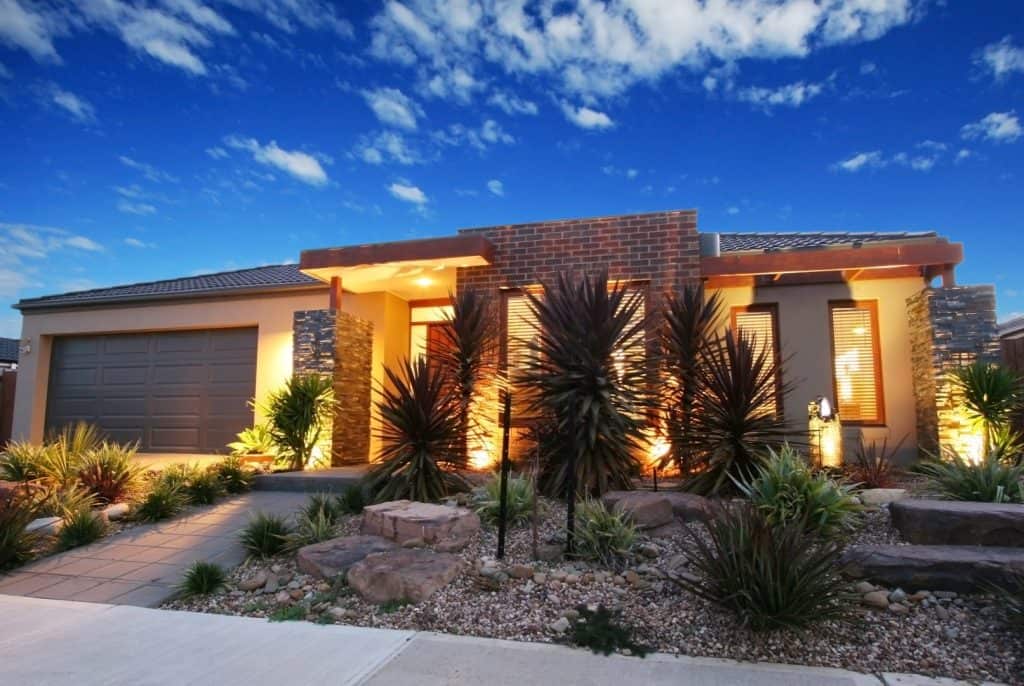 Modern single-story house in CA with brick and stone exterior, large windows, and a double garage—expertly built by a general contractor serving Contra Costa & Alameda County—surrounded by desert landscaping and warm outdoor lights under a deep blue sky.