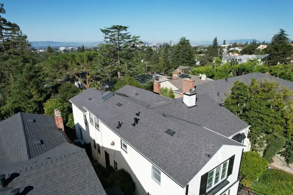 Aerial view of residential houses with gray shingle roofs, surrounded by trees, with a clear blue sky and cityscape in the distance—ideal inspiration for a general contractor in Contra Costa & Alameda County, CA.