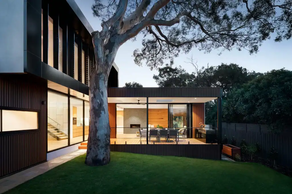 Modern CA house with large windows and an outdoor patio dining area, surrounded by greenery and a tall tree in the backyard at dusk—expertly crafted by a general contractor in Contra Costa & Alameda County.