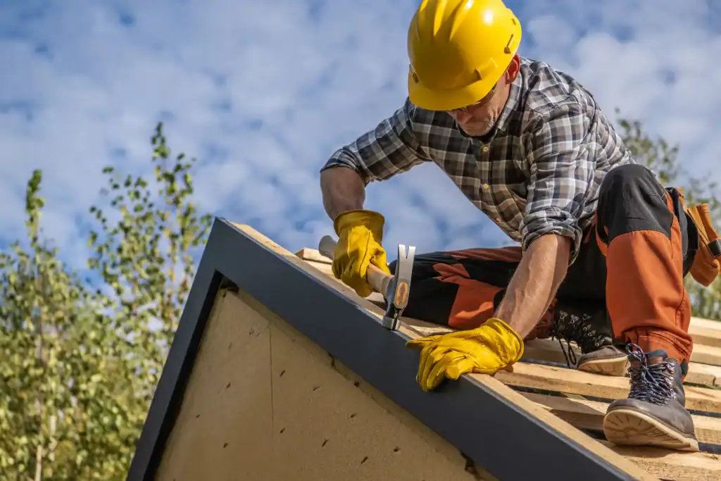 A construction worker wearing a yellow hard hat, gloves, and safety gear hammers a nail into the edge of a roof while working outdoors under a partly cloudy sky. Trees are visible in the background for this general contractor in Contra Costa & Alameda County, CA.