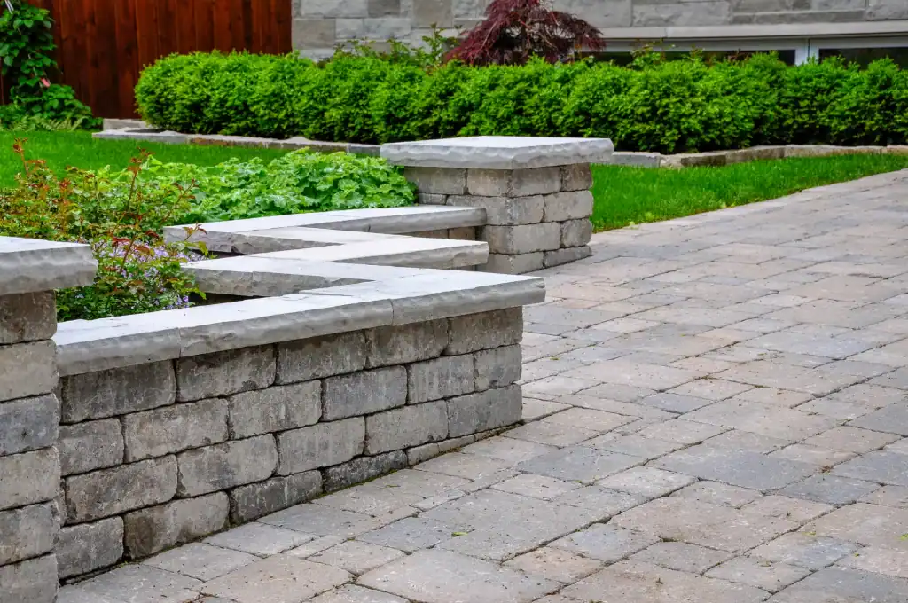 A landscaped yard with a stone retaining wall, neatly trimmed green bushes, a small tree, and a paved walkway—expertly crafted by a general contractor in Contra Costa & Alameda County, CA—creates an inviting garden space.