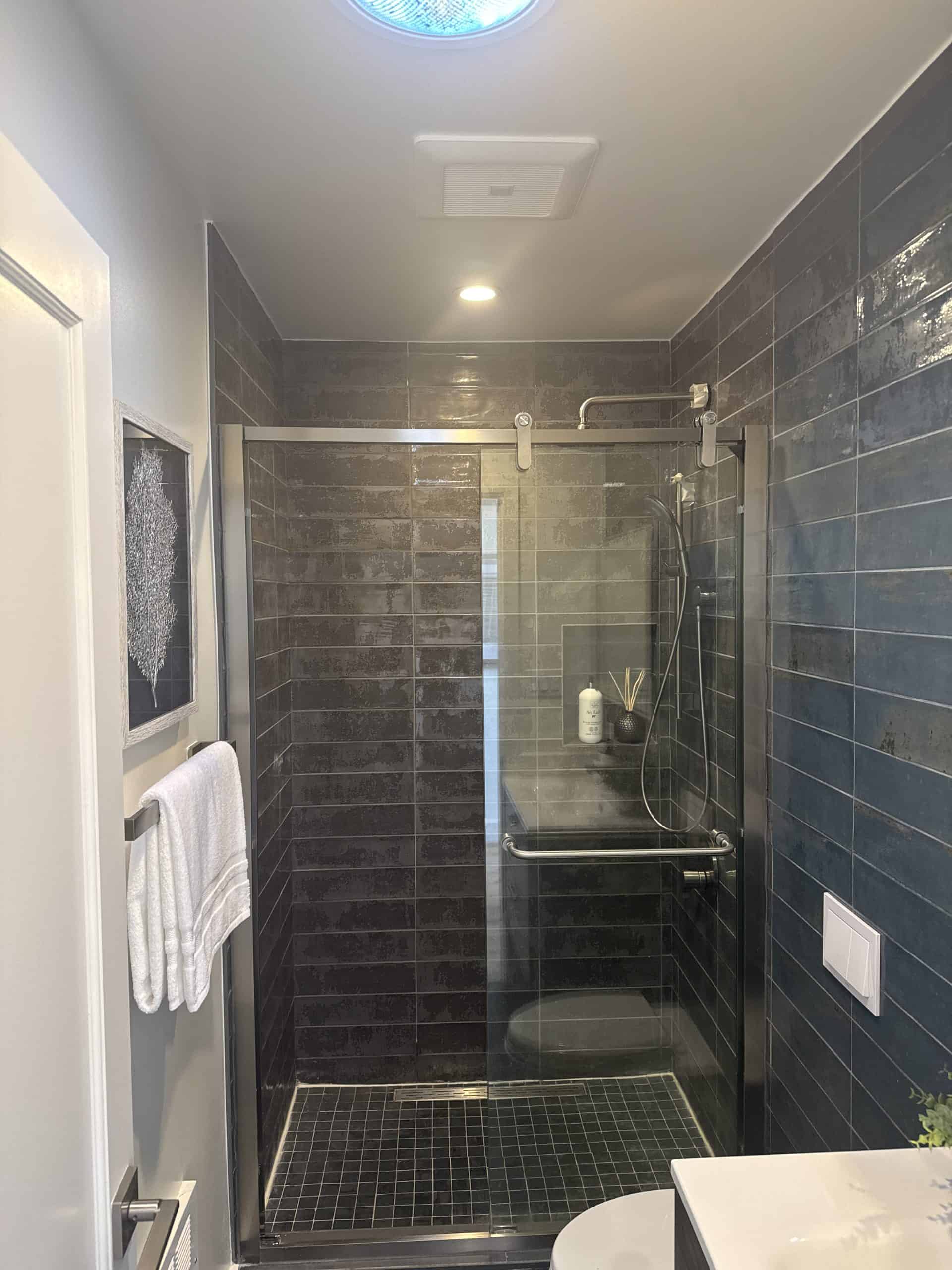 A modern shower with dark gray subway tiles, a glass door, a rainfall showerhead, a towel hanging on the wall, and a small shelf holding toiletries. The space is bright with overhead lighting.
