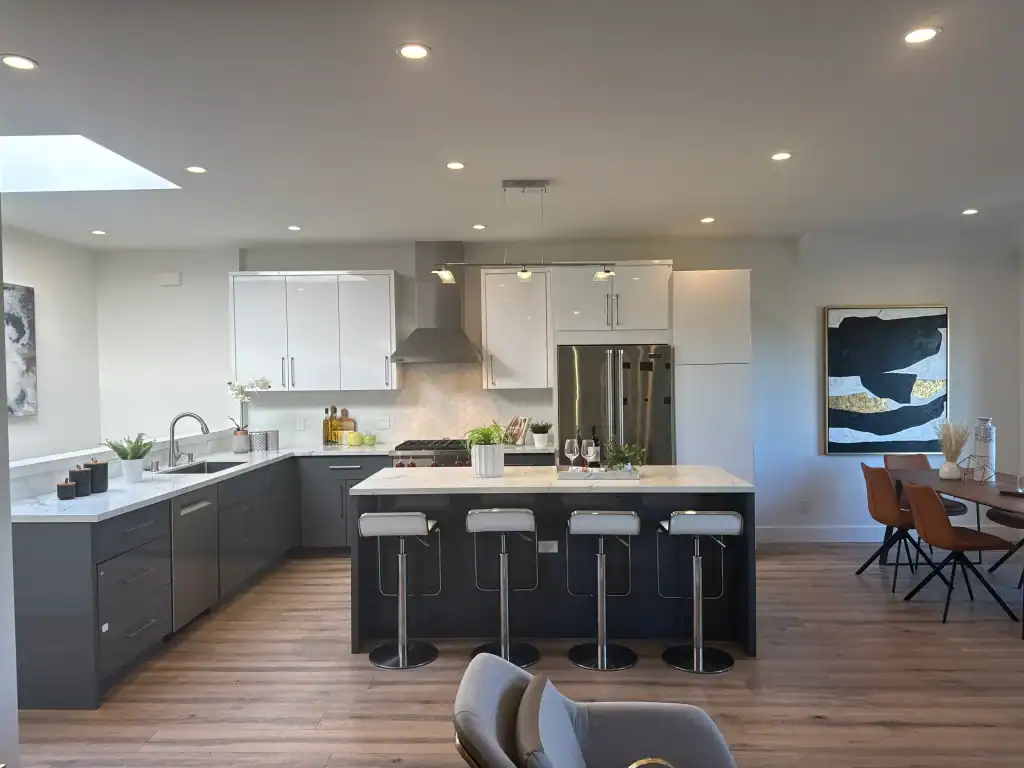 Modern kitchen with white upper cabinets and dark lower cabinets, a large island with four barstools, stainless steel appliances, wood flooring, and a dining area with orange chairs and a contemporary wall art piece.