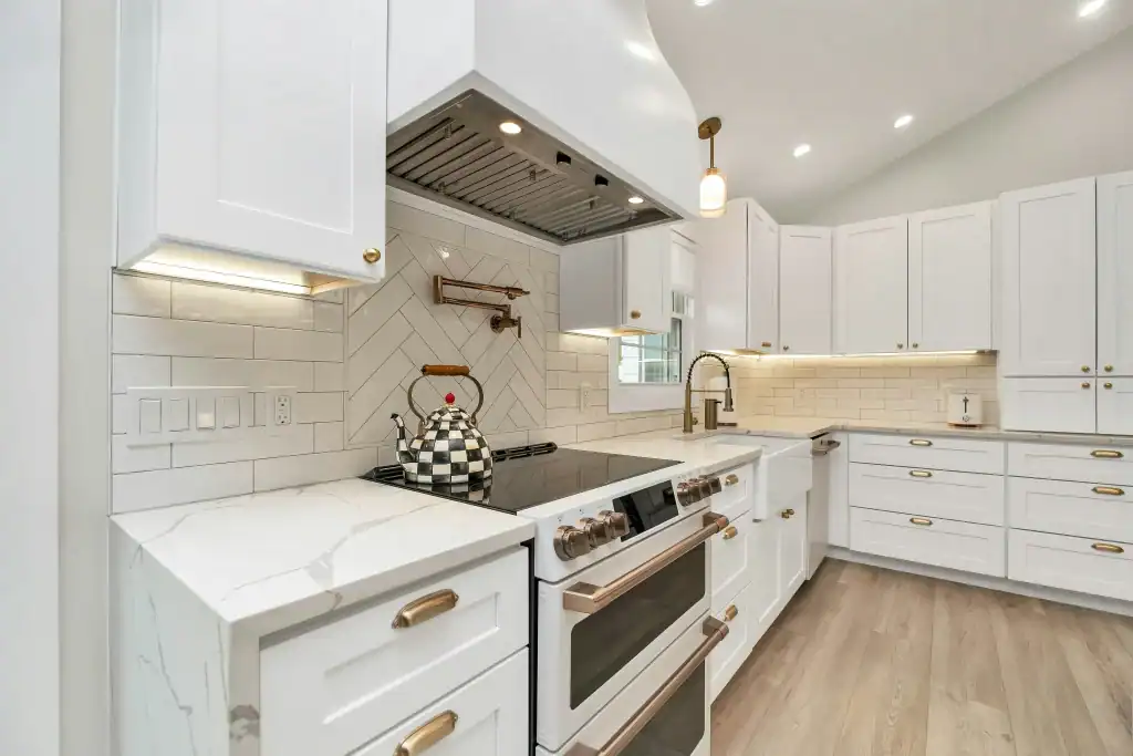 Modern kitchen with white cabinets, gold handles, marble countertops, a double oven, electric stovetop, checkered teapot, herringbone tile backsplash, under-cabinet lighting, and light wood floors.