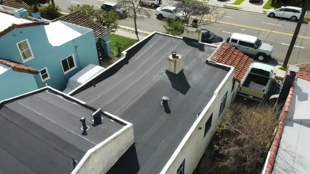 Aerial view of a house with a flat black roof, white walls, and several rooftop vents—perfect for those seeking General Contractor Services in the Bay Area, CA. Surrounded by other houses, parked cars, and lush trees.